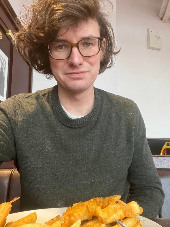 An individual with short, curly hair and glasses is seen wearing a dark-colored top and seated in front of a plain, light-colore