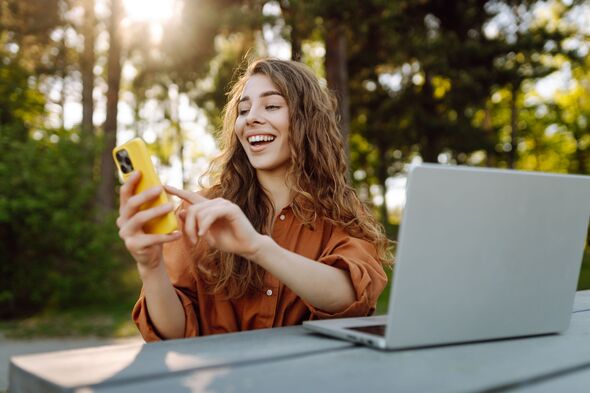 Portrait of a young woman with a phone and laptop at a wooden table outdoors.