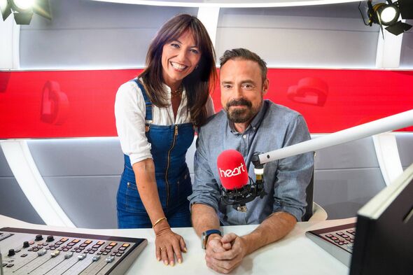 Two individuals, likely professionals, are positioned in a modern radio studio. One person, seated and facing the camera, appear