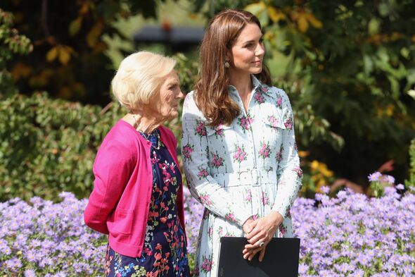 Two women standing in a garden setting, engaged in conversation. One woman is dressed in a pink cardigan, and the other is weari