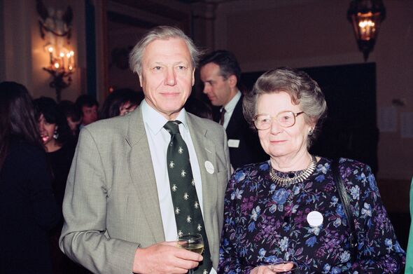 An older man and an older woman stand close to each other at a formal event, with several other people in formal attire visible