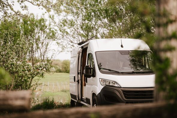 A modern white camper van parked peacefully in a green countryside setting, surrounded by blooming trees and soft natural light.