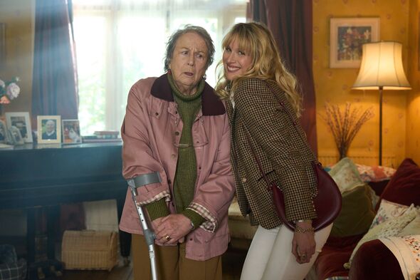 Two individuals are standing in an indoor setting that appears to be a living room. One person is using a cane and appears older