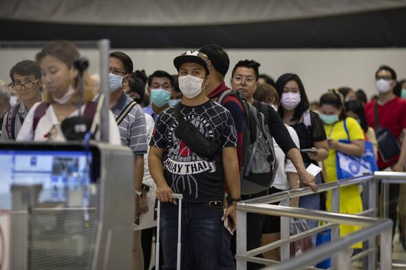 A group of individuals, each wearing protective face masks, are standing in a queue at an airport. Some are carrying backpacks a