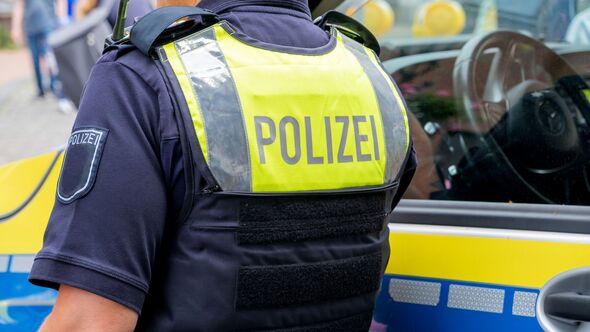 German police officer seen from behind in a yellow vest with Polizei text, standing by patrol car. Suitable as background for breaking news, reports,
