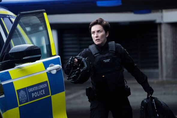 An individual dressed in a black tactical suit is seen standing beside a police vehicle. The individual appears to be holding a