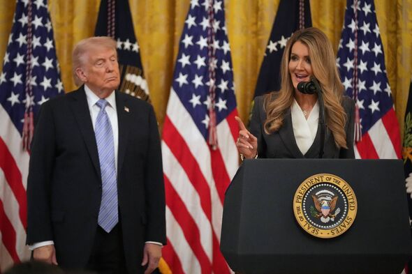 Two individuals are standing at a podium, with one of them speaking into a microphone. Behind them, several American flags are d