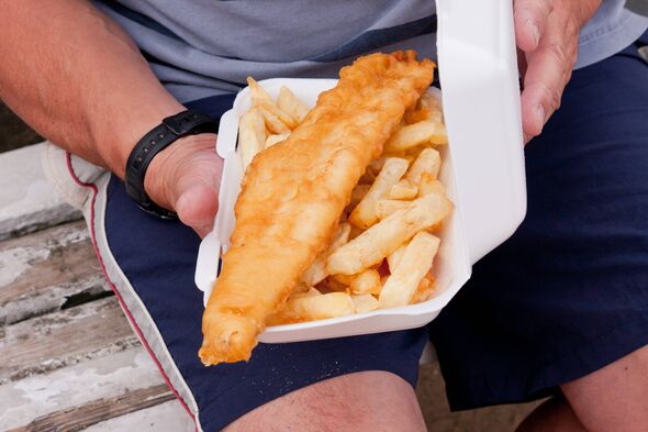 An individual is seated, holding a white cardboard container filled with a portion of fried fish accompanied by a side of golden