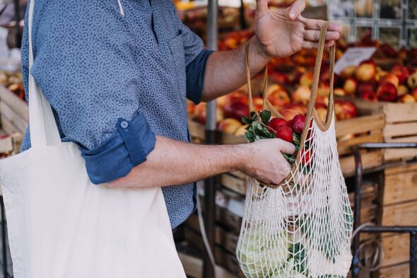Person places bunch of fresh radishes into string mesh bag while holding canvas tote at outdoor market. Buying local produce, open air fruit market, s