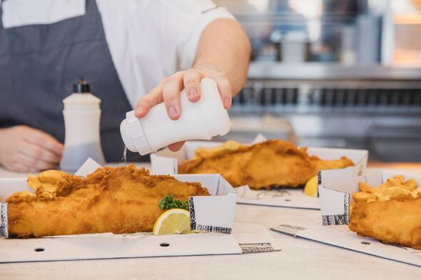 A chef in a white shirt and black apron is seasoning a dish of fried fish, with multiple prepared servings and a lemon slice pla