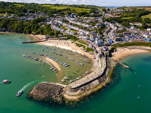 An aerial view of a coastal town featuring a sandy beach, a pier extending into green waters, several boats moored, and a dense