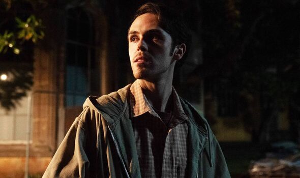 Young man in khaki jacket stands by a large building in the dark