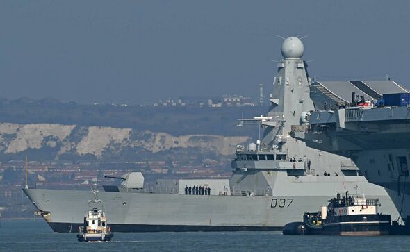 A large gray naval vessel is prominently featured in the image, situated in the foreground with a city skyline visible in the ba