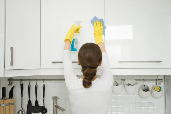 Rear view at an attractive young woman cleaning a surface of white kitchen wall cabinet, wearing rubber protective yellow gloves