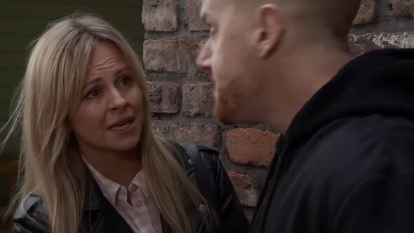 Two individuals engaged in a serious conversation, standing in front of a brick wall. The woman, dressed in a black jacket and w