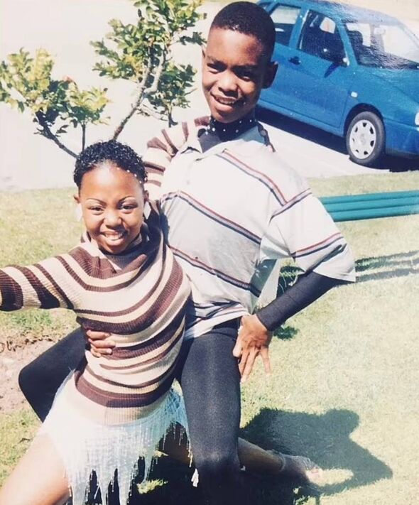 Two individuals are standing close to each other, smiling and posing for a photograph. They are standing next to a young tree wi