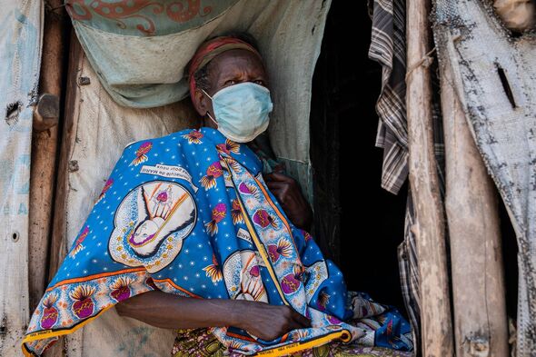 An individual wearing a protective face mask rests within a rudimentary shelter constructed from wooden materials.