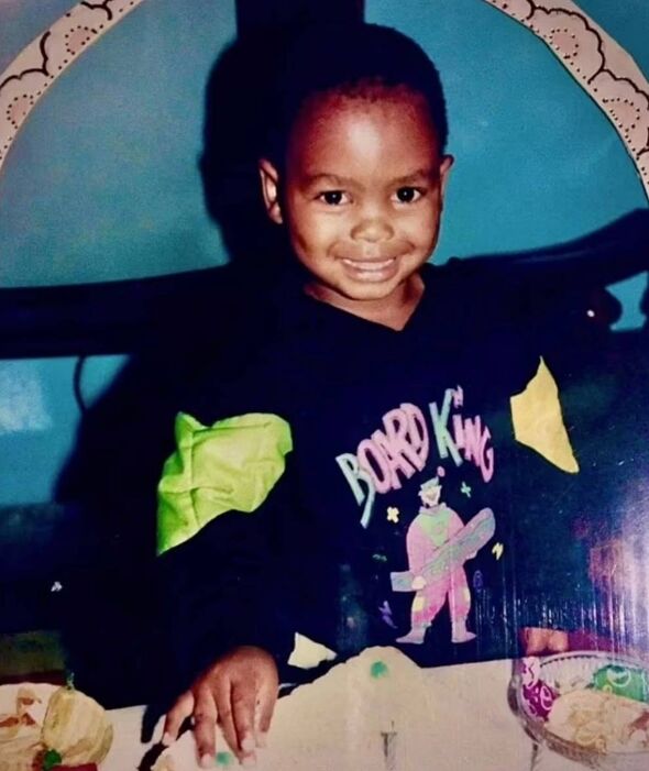 A young child is seated in a chair with a cheerful expression, dressed in a black t-shirt featuring pink text. The background co