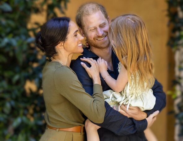 Princess Lilibet is held by Prince Harry as Meghan Markle smiles at her